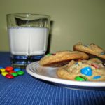 Side view of a few rainbow cookies on a white plate and a glass of milk on a table
