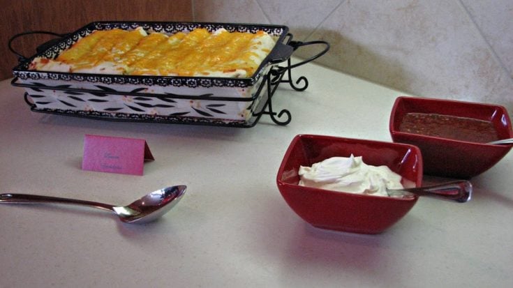 Brunch enchiladas in a serving dish, sour cream in a red bowl, salsa in a red bowl. All on a white table