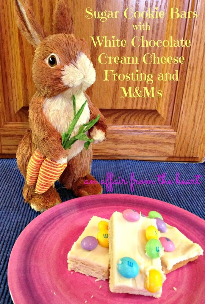 Sugar cookie bars on a pink plate and a plush brown easter bunny on a blue surface. Text "Sugar Cookie Bars with White chocolate cream cheese frosting and M&M's."