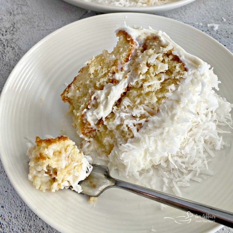A fork with a bite of the COCONUT CREAM POKE CAKE WITH COCONUT WHIPPED CREAM FROSTING next to the slice of cake on a white plate