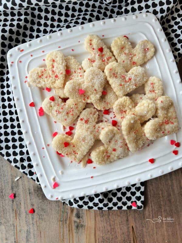 Close up of Heart shaped pie crust cookies with red sprinkles on a white, square dish.