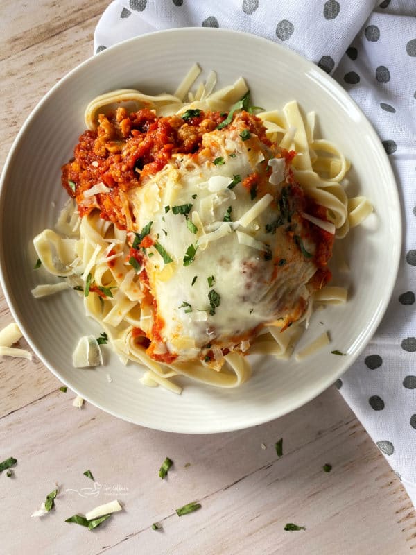 top view of chicken parmesan served over noodles on white plate with fresh parsley and melted cheese