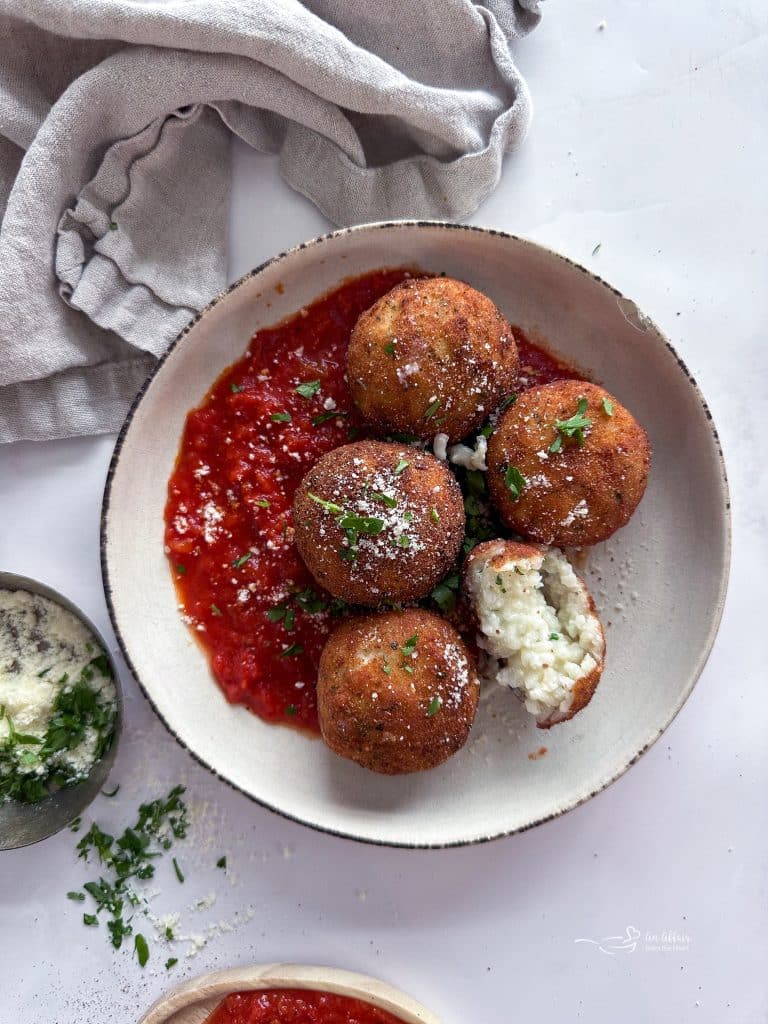 Three Cheese Arancini in a white bowl with tomato sauce. One with a bite out of it.