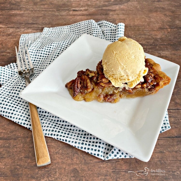 Slice of pecan pie with a scoop of ice cream on top on a white plate with a fork and polka dot napkin on a wood surface