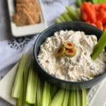 Olive Dip with a celery stick in on a white tray surrounded by celery and carrots