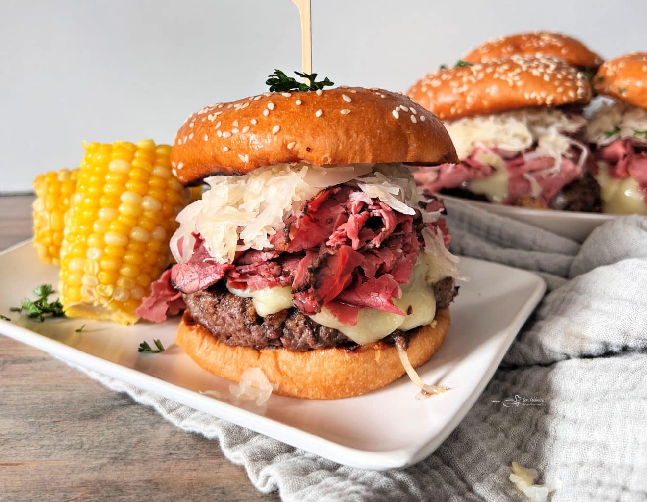 Pastrami Burger with Kraut on a white plate with corn and with burgers behind it