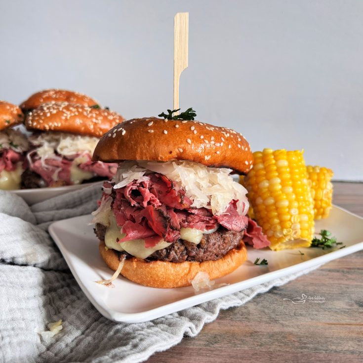 Pastrami Burger with Kraut on a white plate with corn and with burgers behind it