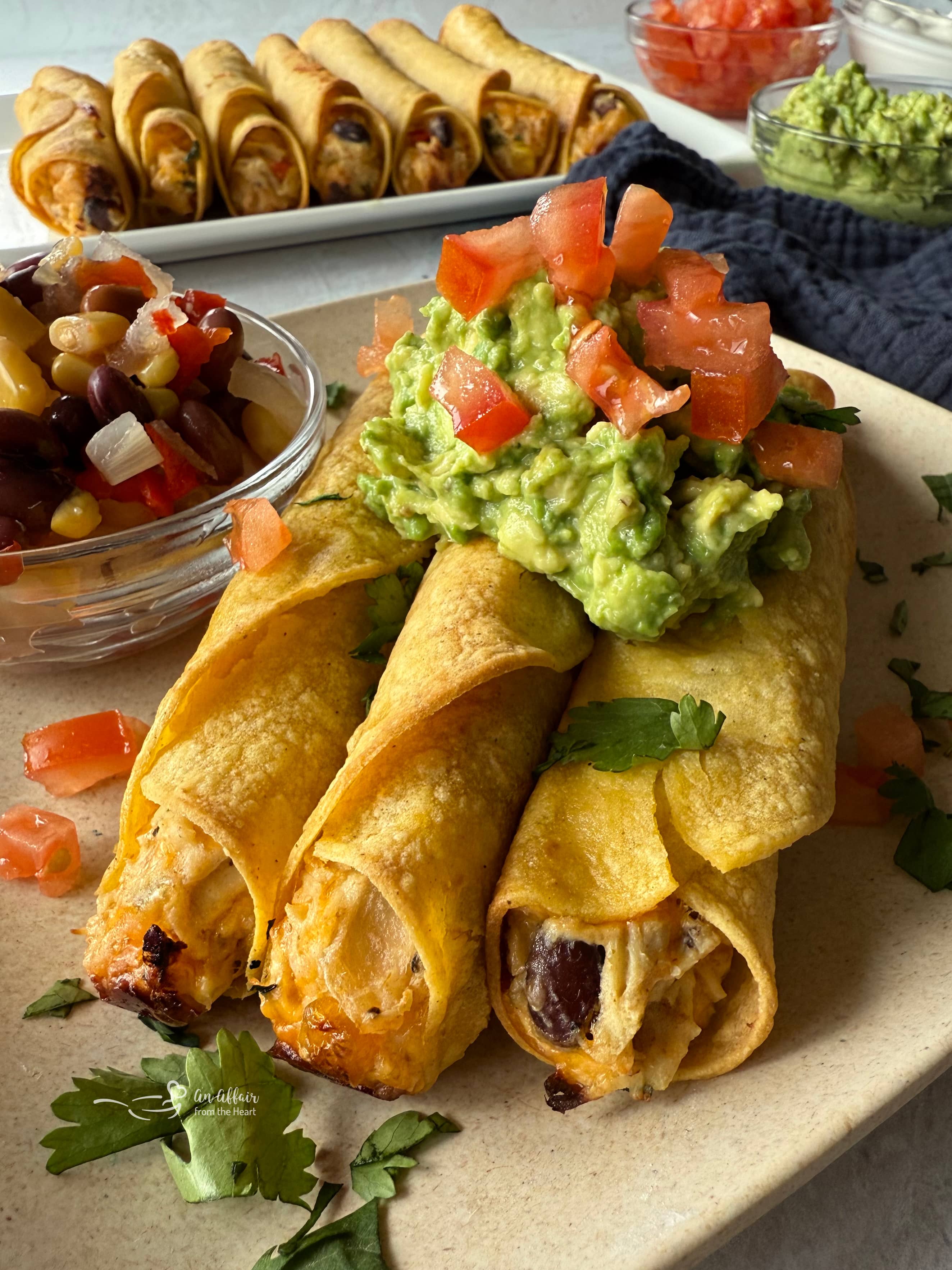 Chicken & Cowboy Caviar Taquitos topped with guacamole and tomatoes on a plate.